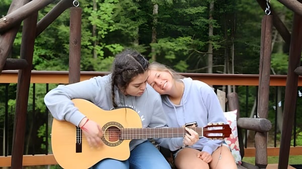 Zwei Schülerinnen der Thornton Academy sitzen auf einer Holzbank im Wald, eine spielt Gitarre, die andere hört zu.