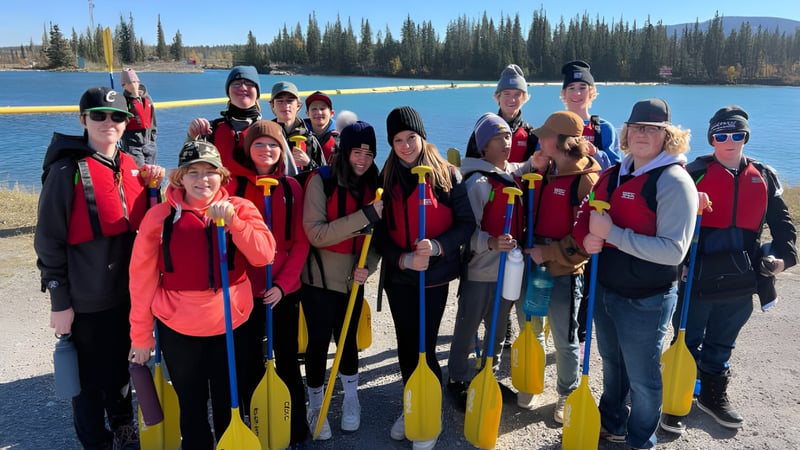Eine Gruppe von Schülern der Three Hills School steht mit roten und gelben Schwimmwesten vor einem See umgeben von Kiefern.