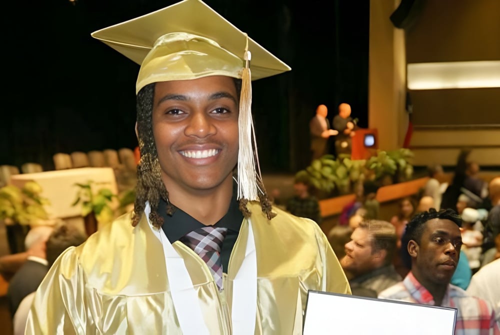 Ein junger Mann in Abschlussrobe hält in der überfüllten Aula ein Diplom auf dem Campus der Three Oaks Senior High.