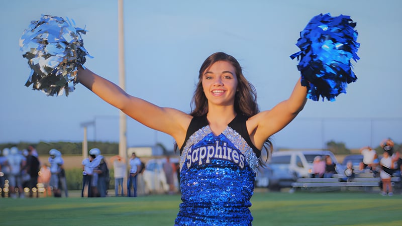 Eine Cheerleaderin der Thunder Ridge High School steht auf dem Sportfeld und winkt mit blauen und silbernen Pompons.