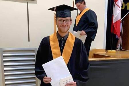 Ein junger Mann steht in Abschlussrobe mit einer Urkunde in einem Raum der Tilbury District High School.