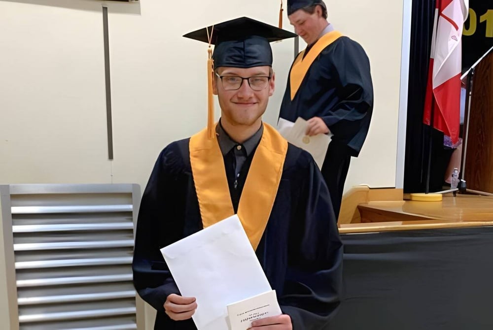 Ein junger Mann steht in Abschlussrobe mit einer Urkunde in einem Raum der Tilbury District High School.