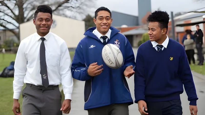 Drei Schüler der Timaru Boys School in Schuluniform mit einem Rugbyball vor einem Schulgebäude und Bäumen.