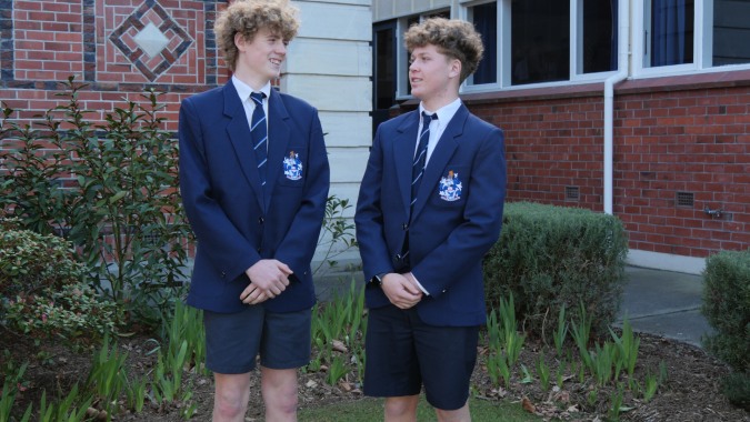 Zwei Schüler der Timaru Boys School stehen in Schuluniform vor einem Backsteingebäude mit Fenstern.