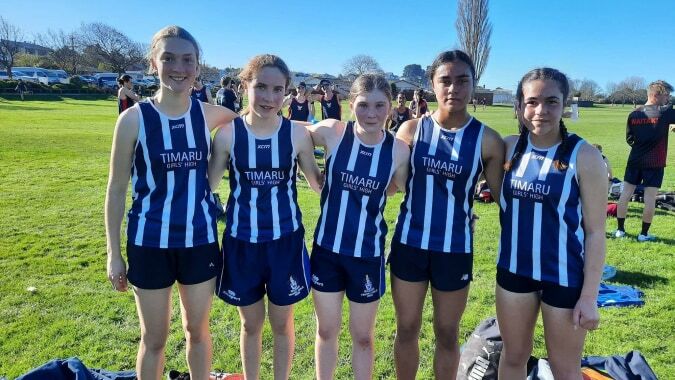 Eine Gruppe junger Athletinnen der Timaru Girls School steht auf einem Grasfeld unter blauem Himmel.