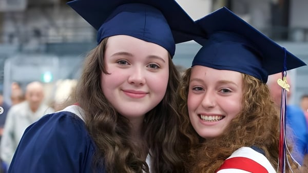 Zwei Schülerinnen in Abschlusskleidung lächeln und posieren gemeinsam vor unscharfem Hintergrund auf dem Campus der Timberline Secondary School.