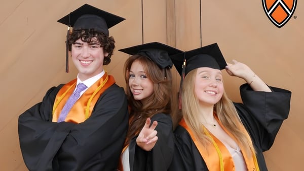 Drei Absolventen der TMI Episcopal School posieren in Abschlusskappen und -roben vor einer Holzwand mit dem Schullogo.