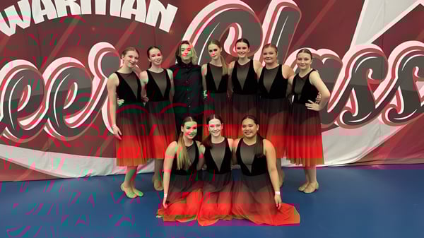 Eine Gruppe junger Schülerinnen der Tommy Douglas Collegiate steht in abgestimmten schwarz-roten Outfits vor einer roten und weißen Grafik als Tanzgruppe.