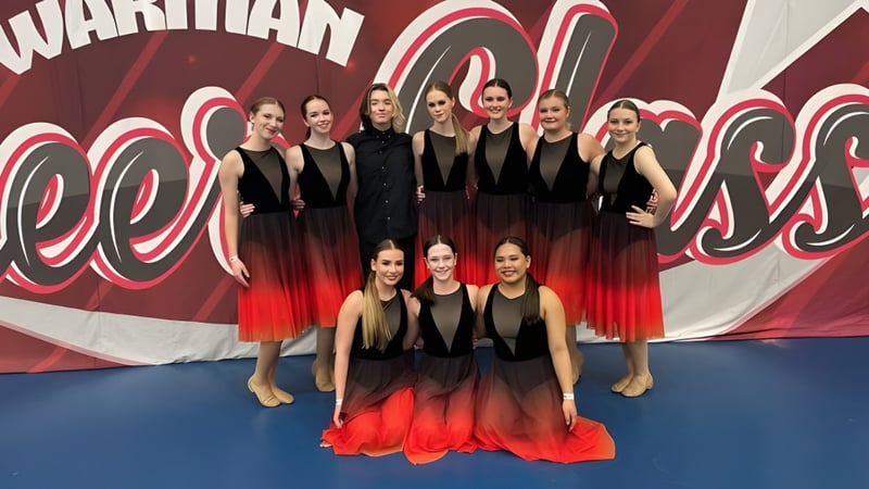 Eine Gruppe junger Schülerinnen der Tommy Douglas Collegiate steht in abgestimmten schwarz-roten Outfits vor einer roten und weißen Grafik als Tanzgruppe.