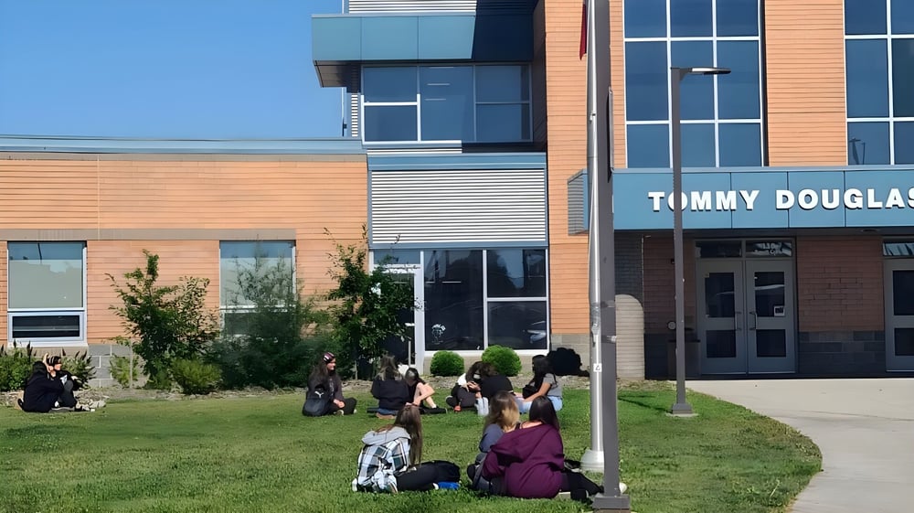 Menschen sitzen auf dem Rasen vor dem modernen Gebäude des Tommy Douglas Collegiate.