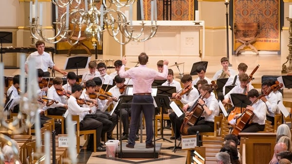 Ein großes Orchester spielt auf der Bühne der Tonbridge School vor dekorativen Kronleuchtern und Architektur.
