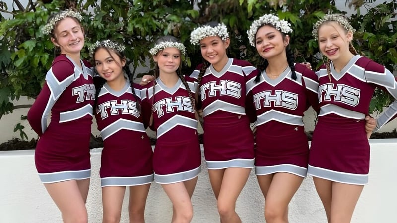 Eine Gruppe Schülerinnen der Torrance High School in Cheerleading-Uniformen mit Blumenkränzen posiert im Freien vor grünem Hintergrund.