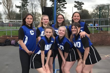 Schülerinnen der Trafalgar School posieren gemeinsam auf dem Sportplatz mit Bäumen und Zaun im Hintergrund.