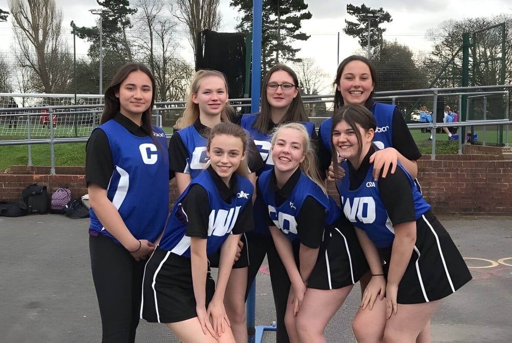 Schülerinnen der Trafalgar School posieren gemeinsam auf dem Sportplatz mit Bäumen und Zaun im Hintergrund.