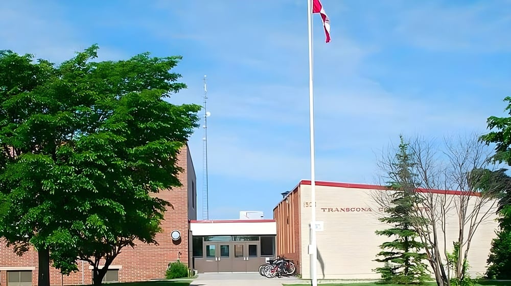 Die Flagge am Fahnenmast weht vor dem Backsteingebäude der Transcona Collegiate und umgeben von grünen Bäumen.