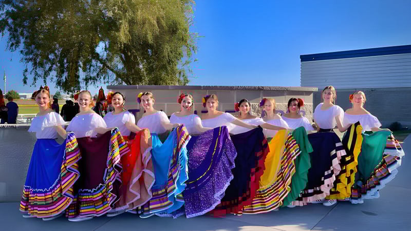 Eine Gruppe von Frauen in traditionellen bunten Kleidern steht vor einem Gebäude der Tri-Cities Prep Catholic High School mit einer Weide im Hintergrund.