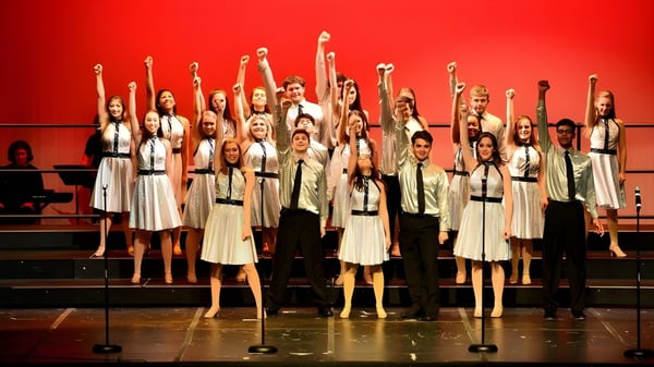 Schüler der Tri-State Christian School stehen in weißen Outfits auf der Bühne und heben die Arme vor rotem Hintergrund.