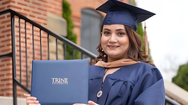 Eine lächelnde Absolventin der Trine University steht vor einem Backsteingebäude und hält ihr Diplom in der Hand.