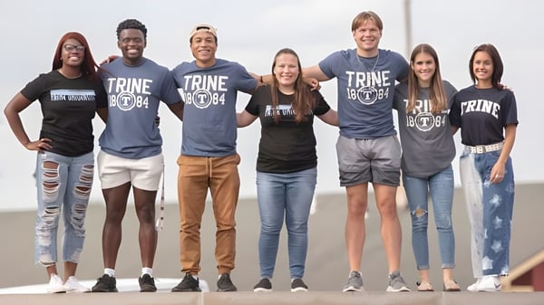 Eine Gruppe Schülerinnen und Schüler steht vor bewölktem Himmel mit Trine University T-Shirts.