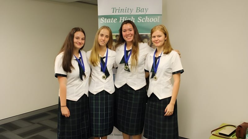 Vier Schülerinnen in Schuluniformen stehen zusammen vor dem Schild der Trinity Bay State High School.