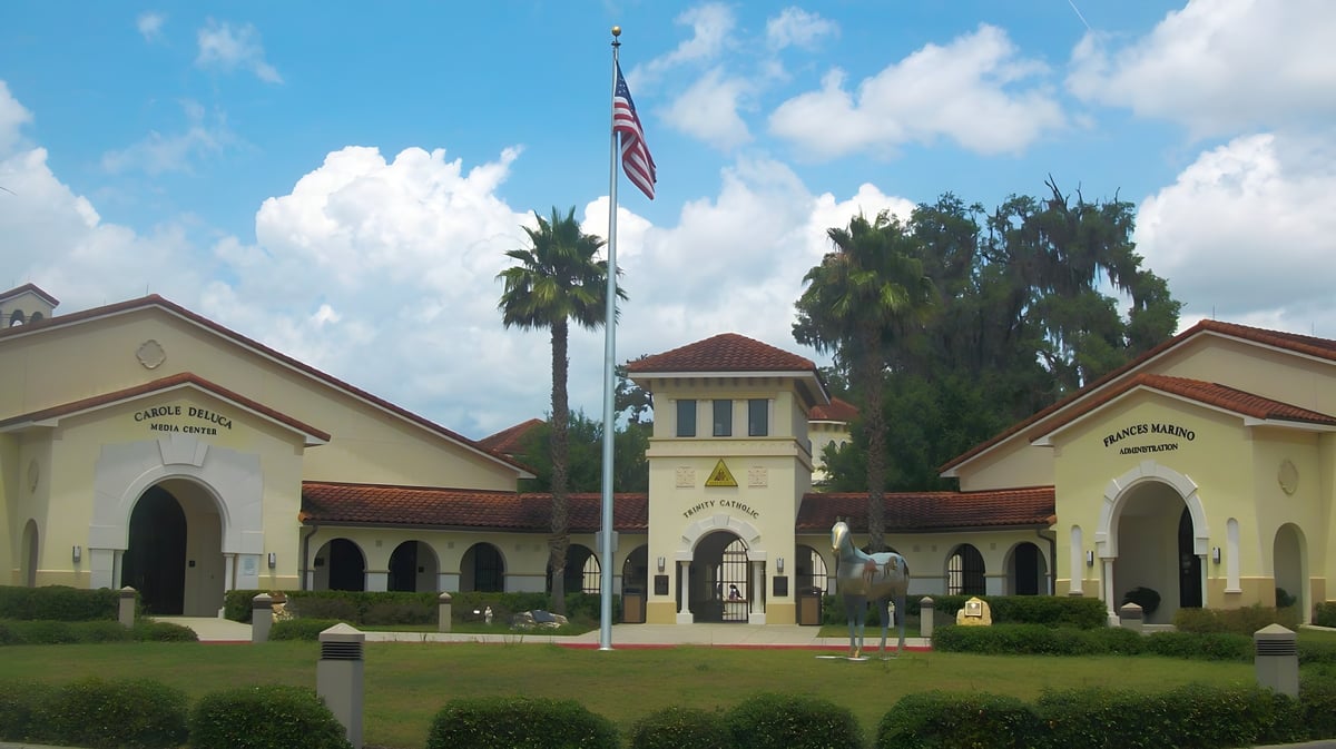 Das Hauptgebäude der Trinity Catholic High School zeigt spanischen Stil mit Turm und amerikanischer Flagge vor Palmen und blauem Himmel.