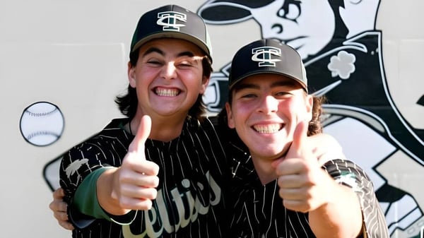 Zwei Schüler der Trinity Catholic High School tragen Baseballkappen und Jerseys und zeigen Daumen hoch vor einem Wandbild mit einer Comicfigur.