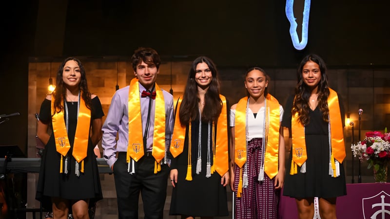 Eine Gruppe von fünf Schülerinnen und Schülern steht in Abschlusskleidung mit Kerzen und Blumen im Hintergrund auf dem Campus der Trinity Christian Academy.