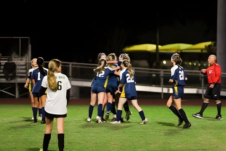 Eine Gruppe weiblicher Fußballspielerinnen der Trinity Preparatory School versammelt sich auf dem Spielfeld bei Nacht mit Schiedsrichter im Hintergrund.