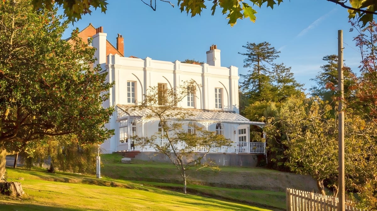 Das weiße herrschaftliche Gebäude der Trinity School steht auf einem grasbewachsenen Hügel mit herbstlich bunt gefärbtem Laub im Hintergrund.