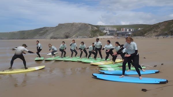 Schülerinnen der Truro High School for Girls stehen mit Neoprenanzügen auf Surfboards am Strand vor Bergen und Gebäuden.