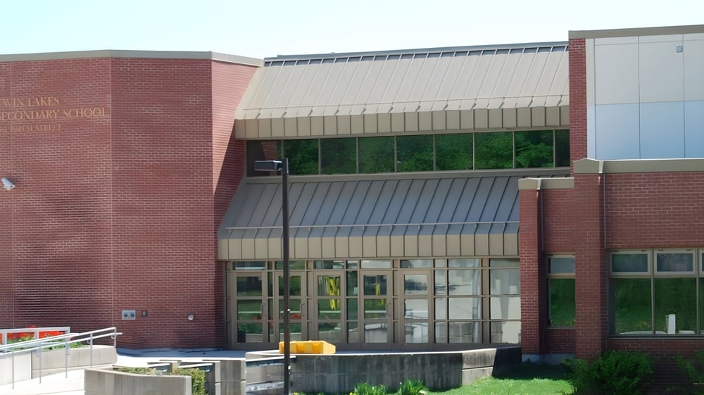 Das Backsteingebäude der Twin Lakes Secondary School mit Glasfenstern und einer Metallüberdachung umgeben von Grünflächen und gepflastertem Vorplatz.