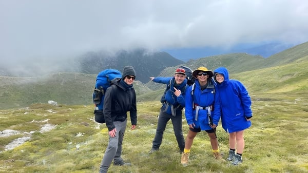 Eine Gruppe von Schülerinnen und Schülern der UC-Lake Ginninderra steht in bunten Regenjacken auf einem nebligen Bergweg mit hohen Gipfeln im Hintergrund.