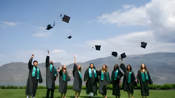 Eine Gruppe von Absolventen der Unisus School wirft ihre Abschlussmützen vor einem See und Bergen in die Luft.