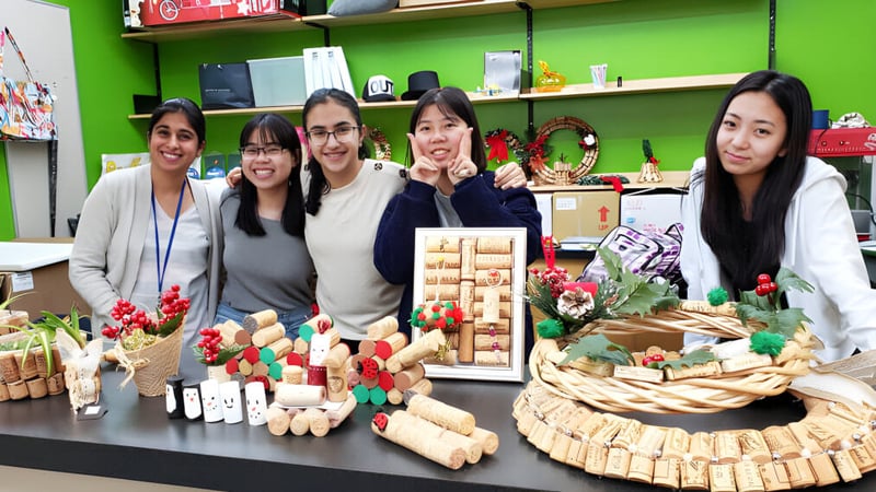 Eine Gruppe Schülerinnen der University Hill Secondary School steht hinter einem Tisch mit verschiedenen weihnachtlichen Bastelarbeiten in einem farbenfrohen Raum.