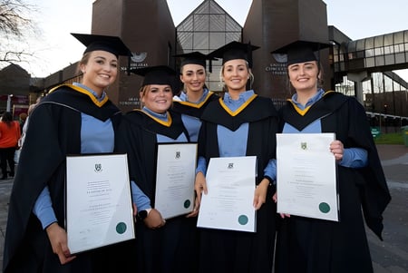Eine Gruppe von fünf Absolventinnen und Absolventen der University of Limerick steht vor dem Universitätsgebäude und hält stolz ihre Diplome.