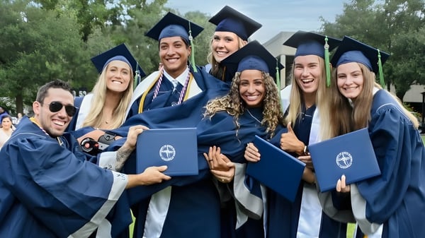 Eine Gruppe von Absolventinnen und Absolventen der University of North Florida steht draußen in blauen Talaren und Hüten umgeben von Bäumen.