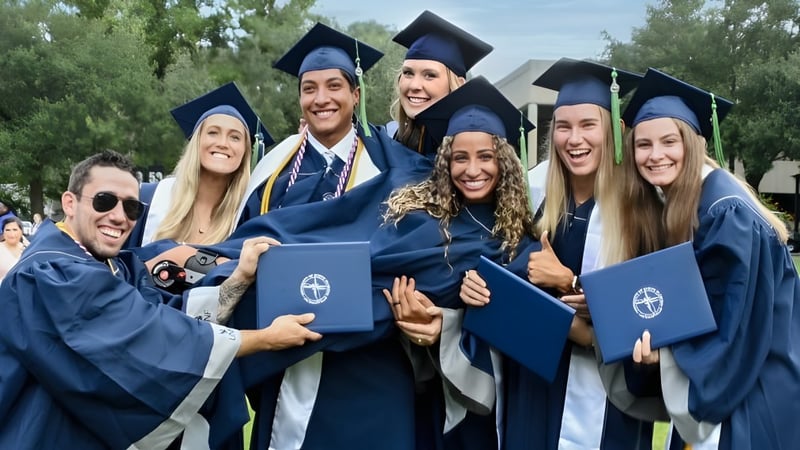 Eine Gruppe von Absolventinnen und Absolventen der University of North Florida steht draußen in blauen Talaren und Hüten umgeben von Bäumen.
