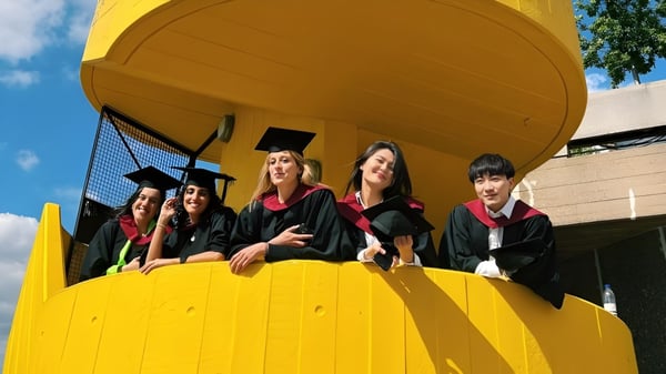 Eine Gruppe von Absolventinnen und Absolventen der University of Westminster steht in roten Roben vor einem gelben Gebäude.