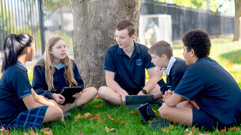 Eine Gruppe Schüler der Unley High School sitzt unter einem Baum und nutzt elektronische Geräte für gemeinsames Lernen.