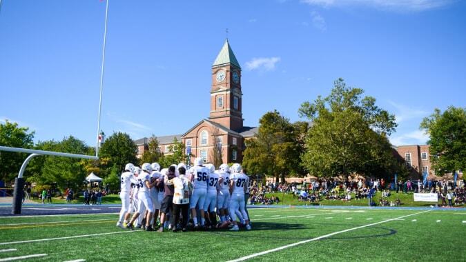 Eine Gruppe Schülerinnen und Schüler in weißen Uniformen versammelt sich auf einem Grasfeld vor einem Backsteingebäude auf dem Campus des Upper Canada College.