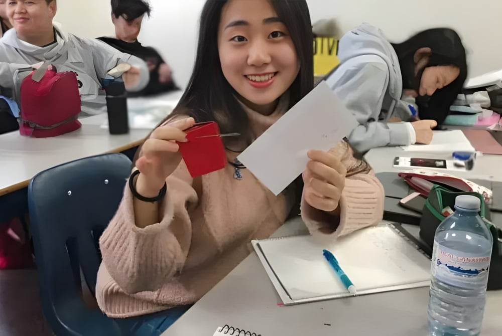 Eine Schülerin der Urban International High School sitzt lächelnd am Schreibtisch und hält einen roten Umschlag in einem Klassenraum.