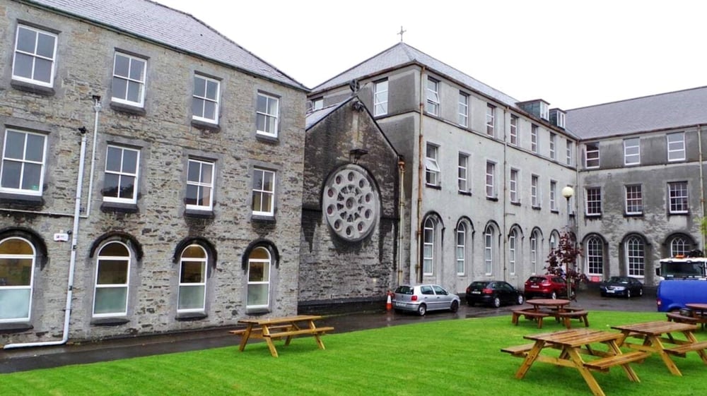 Das historische Gebäude des Ursuline College Sligo mit Rundbogenfenstern und einem großen runden Fenster überblickt den grasbewachsenen Innenhof mit Holzbänken.