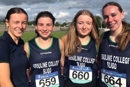 Vier Schülerinnen der Ursuline College Sligo stehen in passenden Sportuniformen auf einem Sportfeld mit bewölktem Himmel.