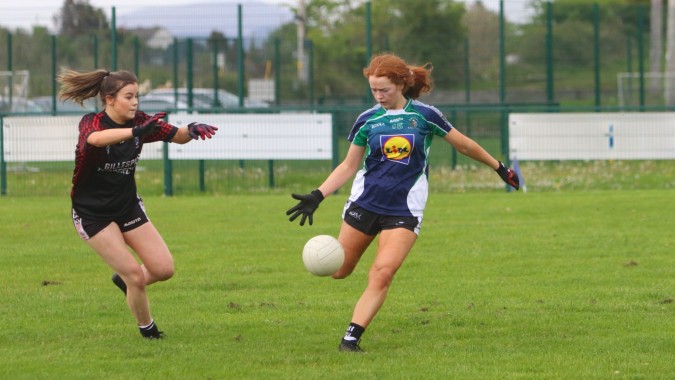 Zwei Schülerinnen des Ursuline College Sligo spielen ein Sportspiel auf einem Rasenfeld mit Wasser und Bäumen im Hintergrund.