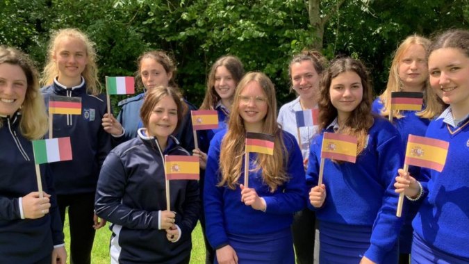 Schülerinnen der Ursuline Secondary School in blauen und gelben Uniformen stehen mit verschiedenen Länderflaggen im grünen Außenbereich.