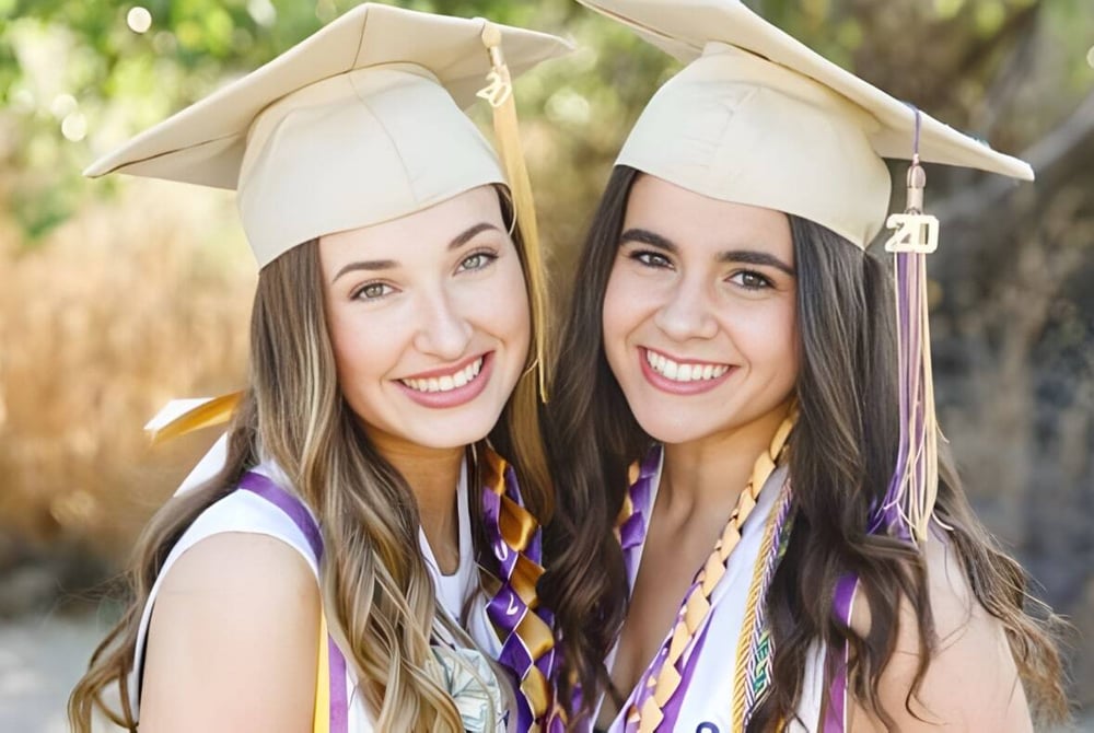 Zwei Schülerinnen der Valencia High School in Abschlussmützen und -roben posieren lachend in einem Feld mit hohem Gras.