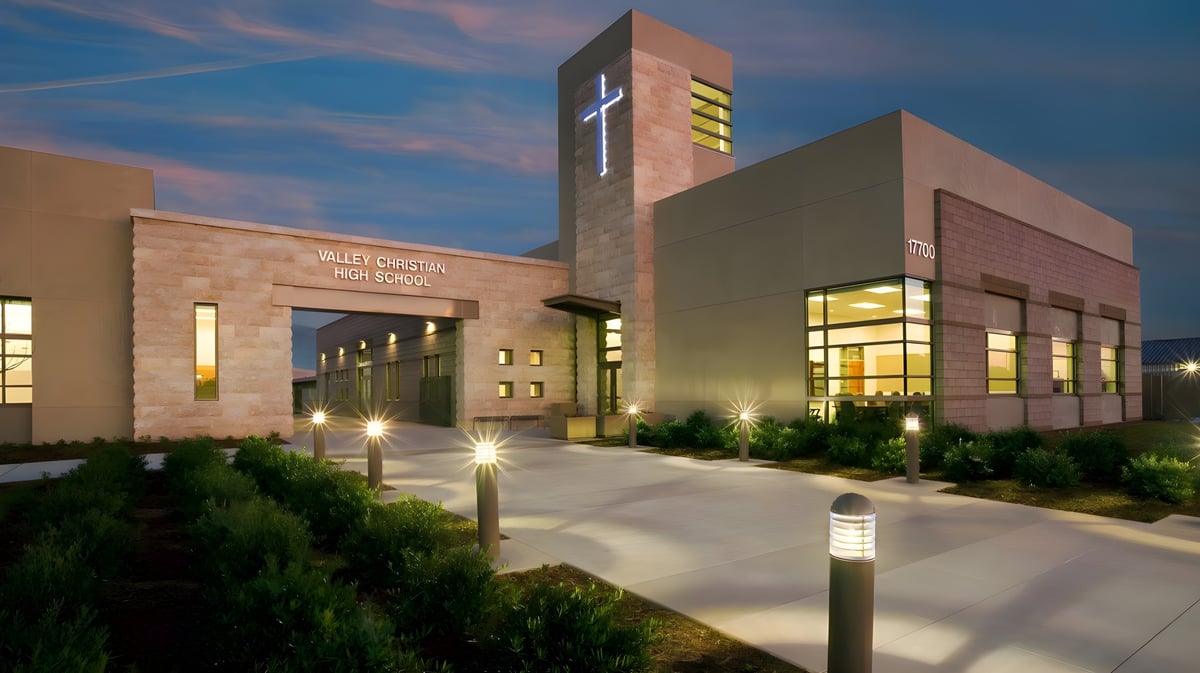 Das mehrstöckige Gebäude der Valley Christian High School mit beleuchtetem Eingang und Türmchen vor einem bunten Abendhimmel.