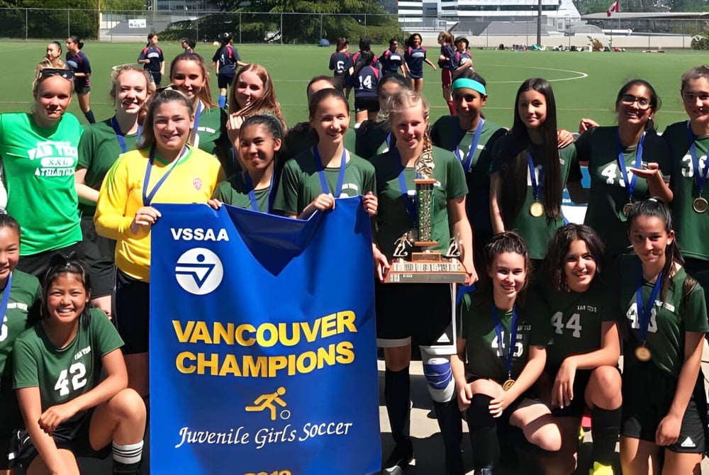 Eine Gruppe junger Fußballspielerinnen der Vancouver Technical Secondary School hält einen Pokal und ein Banner als Jugendmeisterinnen auf dem Spielfeld.