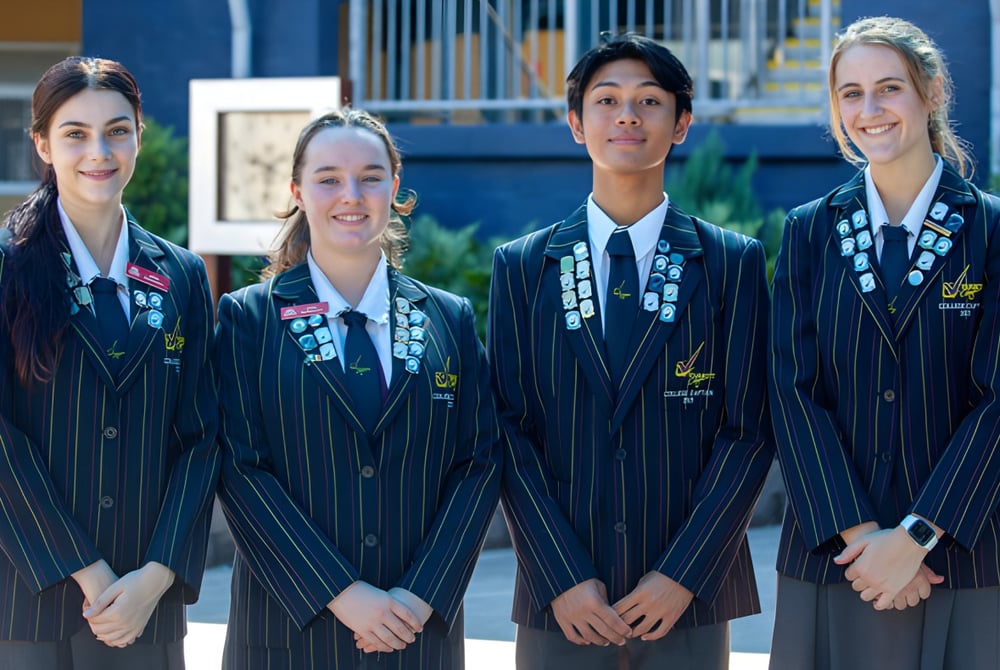 Vier Studentinnen in akademischer Kleidung stehen vor einem Gebäude mit blauer Fassade auf dem Campus des Varsity College.