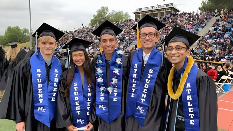 Eine Gruppe Absolventinnen und Absolventen in blauen und gelben Talaren steht bei der Abschlussfeier des Ventura County Community College District vor einem großen Publikum.